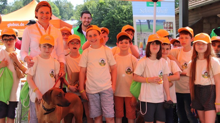 Eine Gruppe von Kindern in beigen T-Shirts und orangenen Mützen steht vor einem GLOBUS-Stand, ein Hund ist vorne zu sehen.