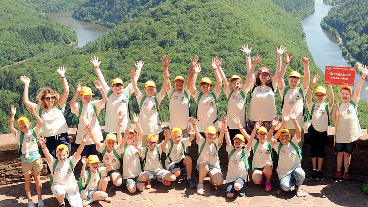 Eine Kindergruppe in weißen T-Shirts posiert mit Globini vor einer grünen Landschaft und hält ein Schild mit Aufschrift „Grundschule Nohfelden“.