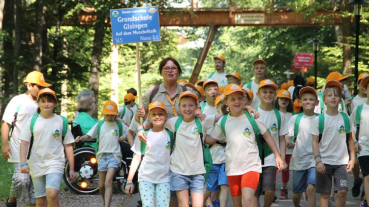 Kindergruppe mit orangenen Kappen und grünen Taschen geht durch den Wald, vorne ein Schild mit „Grundschule Gisingen, Klasse 3.1“.