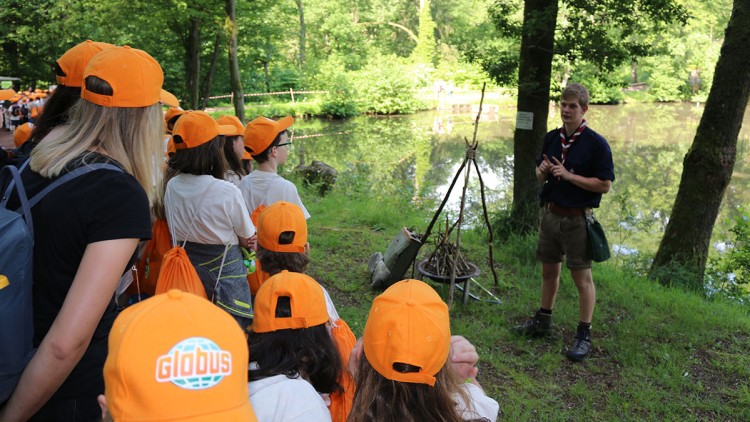 Gruppe von Personen mit orangefarbenen GLOBUS-Kappen steht im Wald nahe eines Sees und hört einer Person zu.