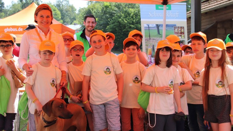 Eine Gruppe von Kindern in weißen T-Shirts mit Globini-Logo und orangenen Kappen steht unter einem GLOBUS-Pavillon, daneben ein Hund.