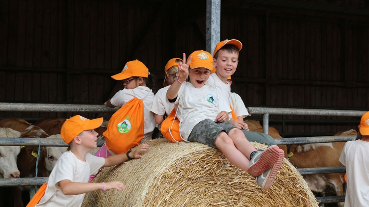 Kinder mit orangefarbenen GLOBUS-Kappen und Rucksäcken sitzen und stehen auf einer Rundballen-Strohrolle vor einem Kuhstall.
