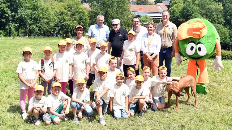 Eine Gruppe von Kindern in weißen T-Shirts und gelben Kappen steht mit Globini, Erwachsenen und einem braunen Hund auf einer Wiese.