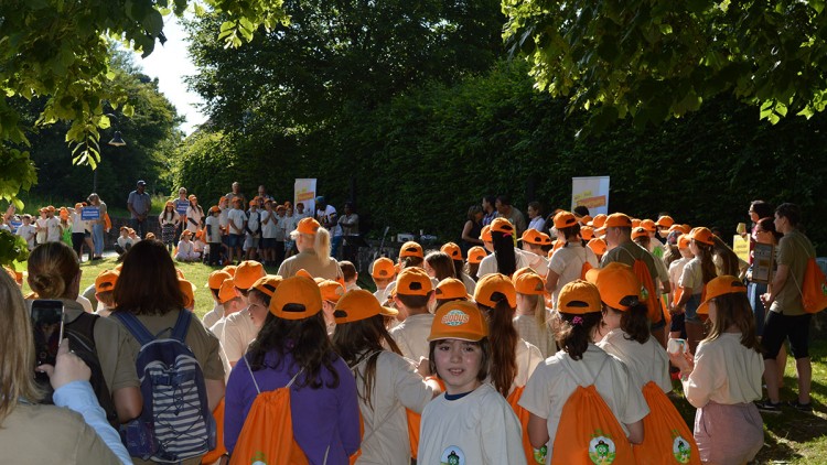 Gruppe von Menschen in orangefarbenen Kappen und beigen T-Shirts auf einer Wiese mit Bäumen, Plakaten und grüner Hecke im Hintergrund.