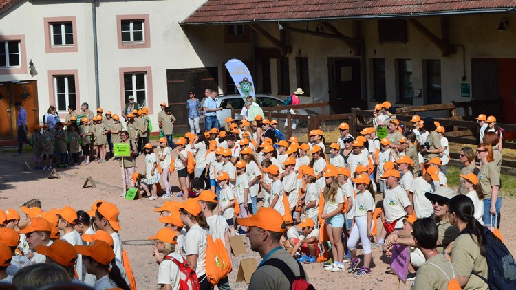 Gruppe von Kindern in beigen T-Shirts und orangefarbenen Kappen steht vor einem Gebäude mit rotem Dach und mehreren Fenstern.