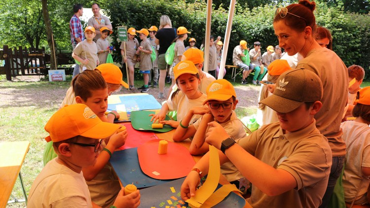 Kinder basteln an einem Tisch im Freien mit bunten Materialien, während andere Kinder in orangefarbenen Mützen im Hintergrund sitzen.