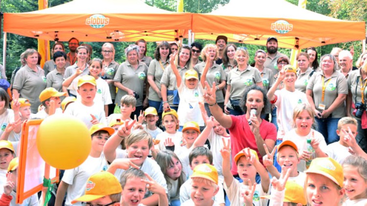 Eine Gruppe von Menschen vor orangen GLOBUS-Pavillons mit Logos, Kinder tragen gelbe Mützen und heben die Arme.