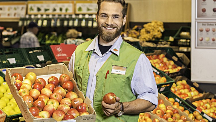 Eine Person in grüner Arbeitskleidung hält eine Kiste mit roten Äpfeln sowie einen einzelnen Apfel in einem Obst- und Gemüsebereich.
