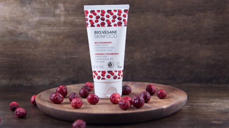 Tube mit der Aufschrift „BIO:VÉGANE SKINFOOD BIO CRANBERRY HANDCREME“ auf einem Holzbrett mit verstreuten Cranberries.