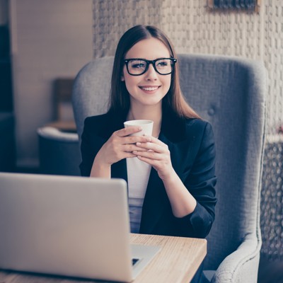 Person in dunklem Blazer sitzt an Holztisch mit Laptop und hält eine Tasse in beiden Händen, im Hintergrund helle Dekoration.