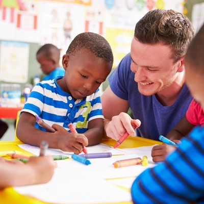 Kinder sitzen an einem gelben Tisch und malen mit Wachsmalstiften auf weißem Papier, im Hintergrund bunte Poster.