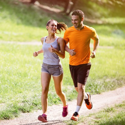Eine Person in grauer Sportkleidung und eine Person in orangem Shirt joggen auf einem Kiesweg durch eine grüne Parklandschaft.