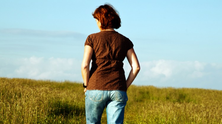 Person in braunem Oberteil und blauer Jeans läuft auf einem grasbewachsenen Weg durch eine Wiese, blauer Himmel im Hintergrund.