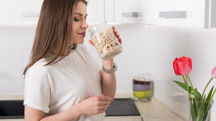 Person in weißem T-Shirt hält ein Glas mit Müsli in einer Küche, daneben eine Schüssel und ein Glas mit Tulpen.