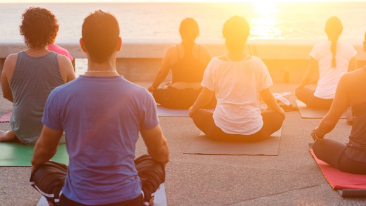 Mehrere Personen sitzen auf Yogamatten im Schneidersitz und blicken bei Sonnenuntergang auf das Wasser.
