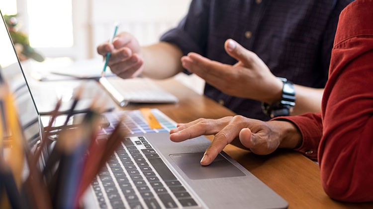 Zwei Personen sitzen an einem Laptop, eine Hand bewegt das Touchpad, die andere hält einen Stift, auf dem Tisch liegen Stifte.