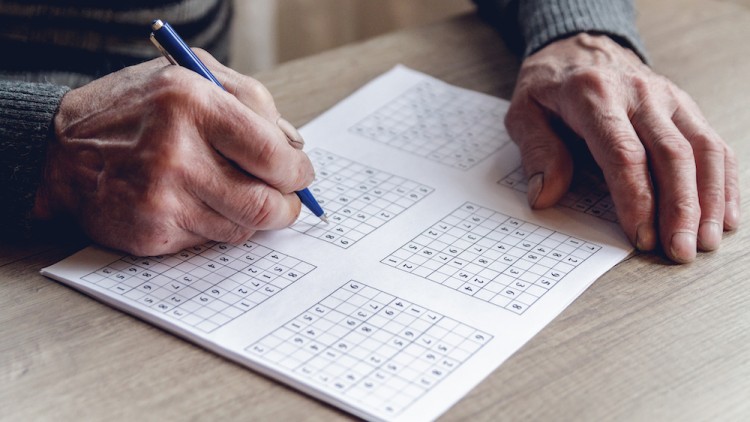 Eine Person schreibt mit einem blauen Kugelschreiber Zahlen in ein gedrucktes Sudoku auf einem Holztisch.