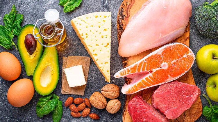 Verschiedene Lebensmittel wie Avocado, Eier, Käse, Nüsse, Fleisch, Fisch, Spinat, Öl, Brokkoli und Äpfel auf grauem Untergrund.