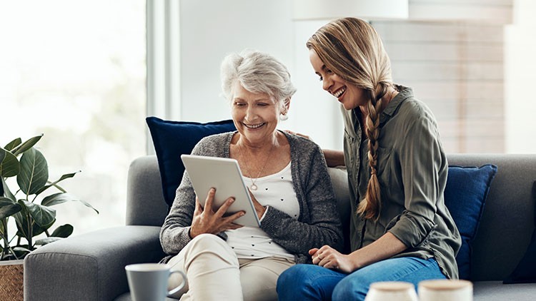 Eine ältere Person und eine jüngere Person sitzen auf einem Sofa, beide schauen gemeinsam auf ein Tablet, umgeben von Kissen und Pflanzen.