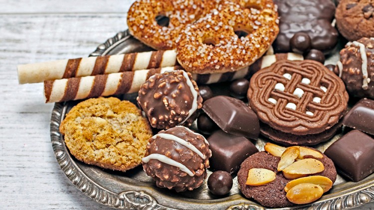 Eine silberne Platte mit Keksen, Schokopralinen, Waffelröllchen und Gebäck mit Mandeln und Zuckerstreuseln auf weißem Untergrund.