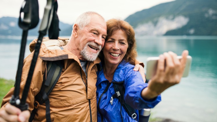 Zwei Personen in Outdoor-Kleidung machen mit einem Smartphone ein Selfie, im Hintergrund ein See und Berge.