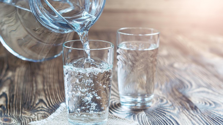 Wasser wird aus einem Glaskrug in ein Wasserglas gegossen, daneben steht ein weiteres gefülltes Glas auf einem Holztisch.