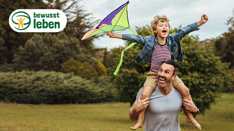 Kind sitzt auf den Schultern einer Person, hält einen bunten Drachen, links oben Logo mit Text „bewusst leben“.