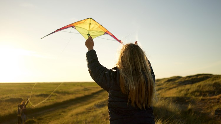 Person mit langen Haaren hält einen bunt dreieckigen Drachen in die Luft, weite Graslandschaft bei Sonnenuntergang.