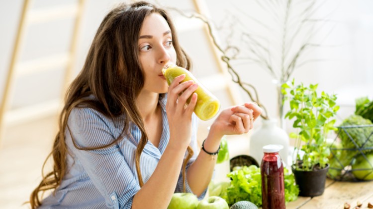 Person in heller Bluse trinkt aus einem Glas mit grünem Smoothie, umgeben von frischem Gemüse, Kräutern und einer roten Flasche.