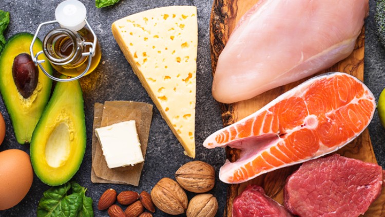 Avocadohälften, Käse, Lachs, Hähnchen, Rindfleisch, Nüsse, Mandeln, Eier, Butter, Basilikum und Olivenöl auf grauem Untergrund.