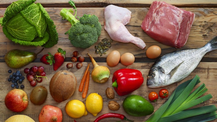 Verschiedene Lebensmittel wie Fisch, Fleisch, Gemüse, Obst, Nüsse und Eier liegen auf einem rustikalen Holztisch.