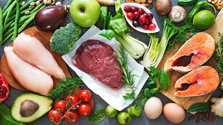 Verschiedene frische Lebensmittel wie Fleisch, Fisch, Gemüse, Obst, Eier und Kräuter auf einem grauen Untergrund.