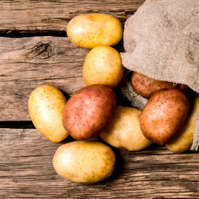 Verschiedene rohe Kartoffeln, gelb- und rotbraun, liegen teils aus einem groben Jutebeutel auf einer rustikalen Holzoberfläche.
