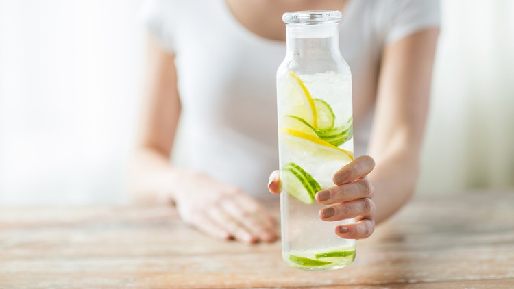Eine Hand hält eine Glasflasche mit Wasser, Zitronenscheiben, Gurkenscheiben und Eiswürfeln auf einem Holztisch.