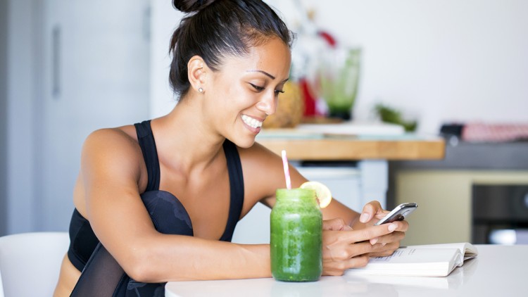 Person in Sportkleidung sitzt an einem Tisch mit einem grünen Smoothie im Glas, einem Buch und einem Smartphone in der Hand.