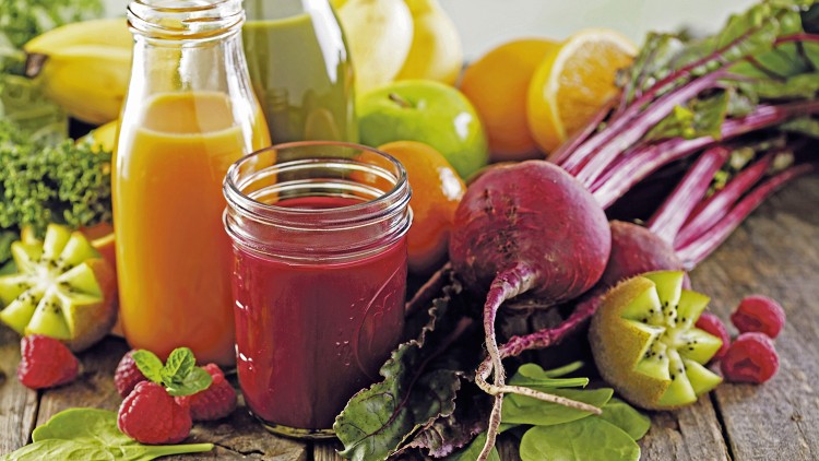 Zwei Gläser Saft aus orangenen und roten Zutaten stehen auf einem Holztisch, umgeben von Obst, Gemüse und Blattspinat.