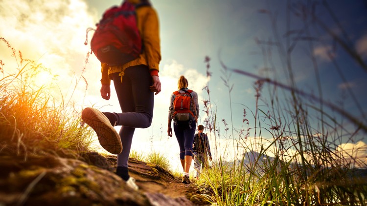 Drei Personen mit Rucksäcken wandern auf einem Pfad durch eine mit Gras bewachsene Landschaft bei Sonnenuntergang.