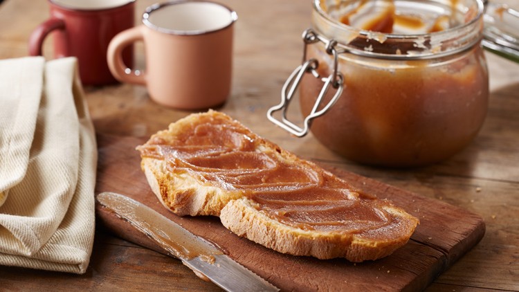 Scheibe Brot mit Aufstrich liegt auf einem Holzbrett, daneben ein Messer, ein Glas mit Aufstrich und zwei Tassen im Hintergrund.