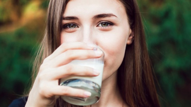 Eine Person mit langen braunen Haaren hält mit der rechten Hand ein durchsichtiges Glas, im Hintergrund sind grüne Pflanzen.