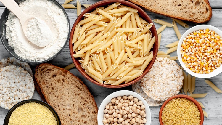 Schale mit ungekochten Penne-Nudeln, daneben Brot, Mehl, Puffreis, Kichererbsen, Maiskörner, Couscous und Bulgur auf Holzuntergrund.