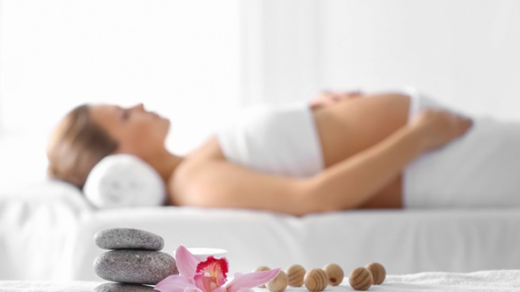 Person liegt in heller Umgebung auf einer Liege, im Vordergrund liegen graue Steine, Holz-Massagekugeln und eine pinke Orchidee.