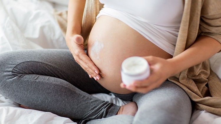 Person in heller Kleidung sitzt im Schneidersitz und cremt einen runden Bauch mit einer weißen Creme aus einem offenen Tiegel ein.