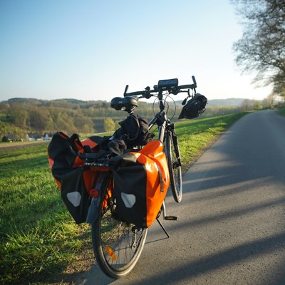 Ein Fahrrad mit orangefarbenen Gepäcktaschen steht auf einem Weg, umgeben von grüner Wiese und hügeliger Landschaft.