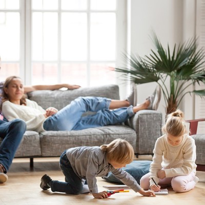 Zwei Kinder malen am Boden mit Stiften, während zwei Erwachsene entspannt auf einem grauen Sofa sitzen, dahinter eine Pflanze.