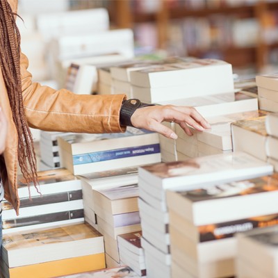 Eine Person in brauner Jacke greift nach einem Taschenbuch auf einem Tisch voller gestapelter Bücher in einem Buchladen.