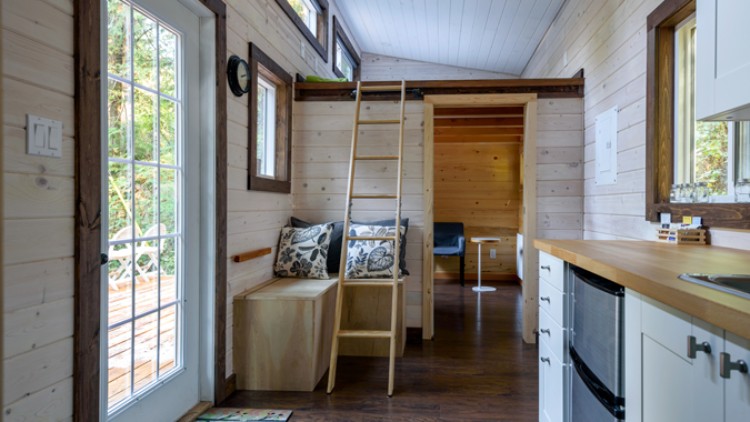 Holzinterieur eines kleinen Hauses mit Leiter zu einem Hochbett, Küchenzeile rechts und Glastür links mit Blick nach draußen.