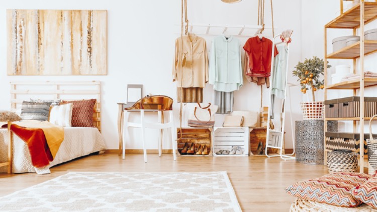 Ein helles Schlafzimmer mit Bett, Holzregalen, aufgehängter Kleidung, Schreibtisch, Teppich und dekorativen Kissen.