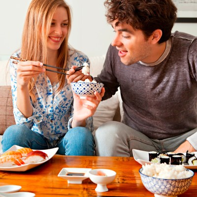 Zwei Personen sitzen an einem Tisch mit Sushi, Reis und Sojasoße, dabei wird eine Reisschale mit Stäbchen gehalten.