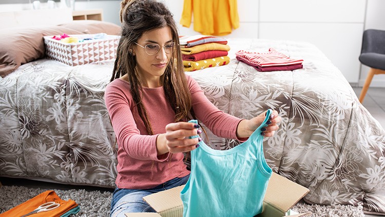 Person sitzt auf einem Teppich vor einem Bett und hält ein türkisfarbenes Oberteil über einem offenen Karton hoch.