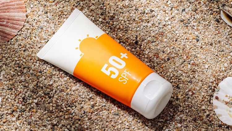 Orange-weiße Sonnencreme-Tube mit Aufdruck „50+ SPF“ liegt auf hellem Sand mit Muscheln.
